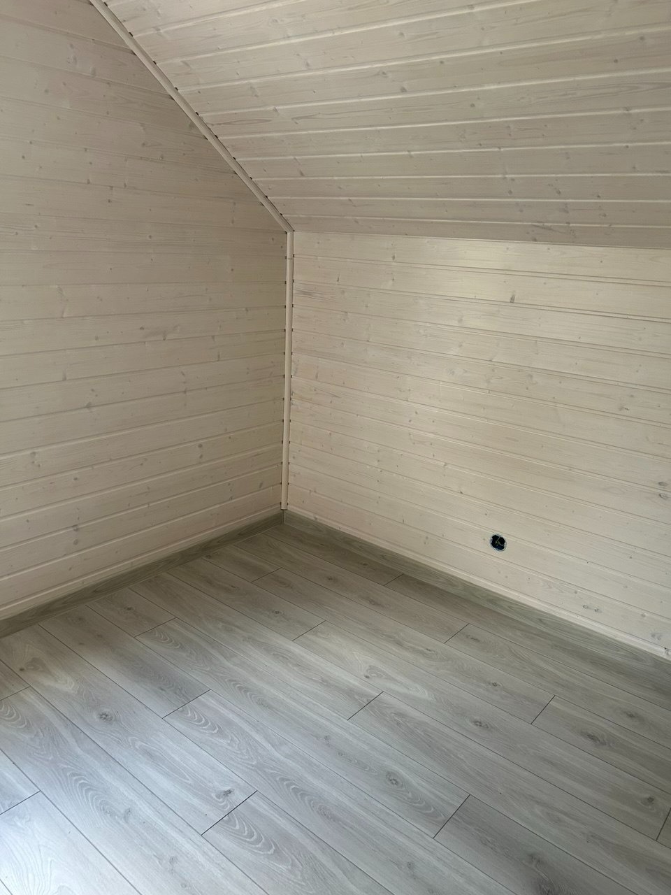 Jasne panele podłogowe i boazeria w odcieniach bieli tworzą minimalistyczne wnętrze poddasza. Widoczne gniazdko elektryczne. Ukośny sufit.