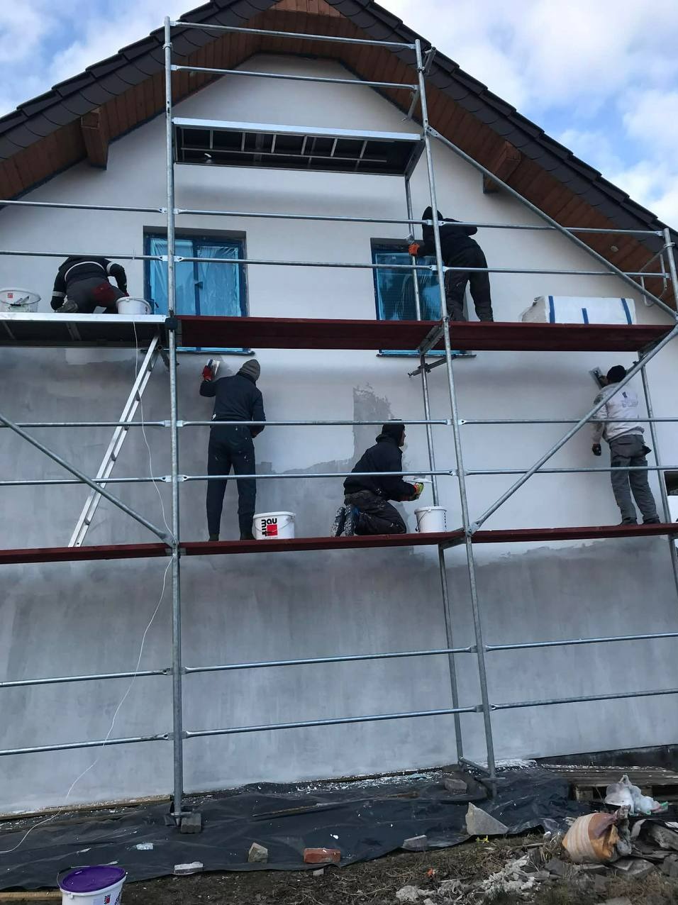 Ekipa na rusztowaniu tynkuje elewację domu jednorodzinnego. Okna zabezpieczone folią. Widoczne wiadra z zaprawą i narzędzia. Częściowo otynkowana ściana.