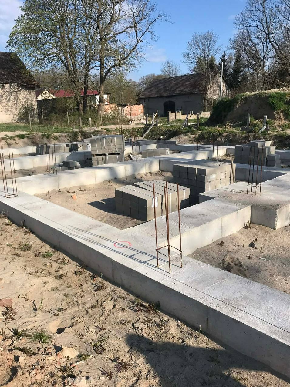 Fundamenty domu jednorodzinnego w Karlino, zbrojenie i bloczki betonowe, widok z poziomu gruntu, w tle budynki gospodarcze i drzewa.