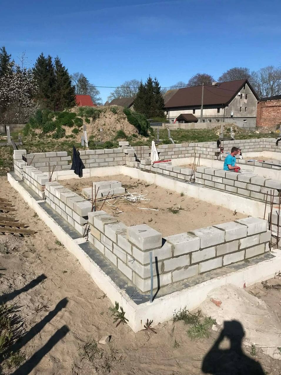 Wznoszenie fundamentu z bloczków betonowych na piasku, widoczne zbrojenia i pracownik w tle. Budowa domu w słoneczny dzień.