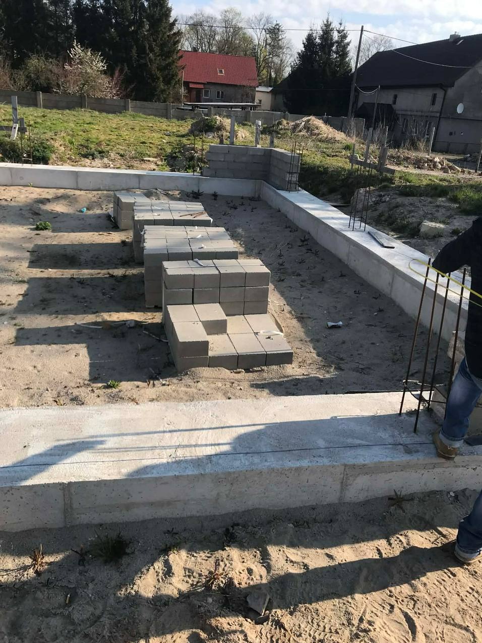 Wznoszenie fundamentu z bloczków betonowych na piaszczystym podłożu. Widoczne zbrojenia i częściowo ukończona ściana fundamentowa. W tle dom z czerwoną dachówką.