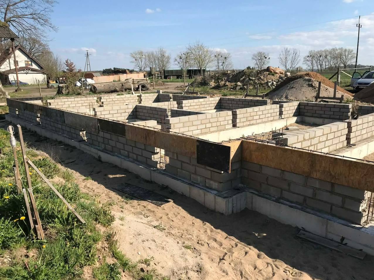 Wieniec fundamentowy z szalunkiem i zbrojeniem na budowie domu jednorodzinnego. Widoczne bloczki betonowe i stalowe pręty. Tło: teren budowy z zielenią i budynkami.