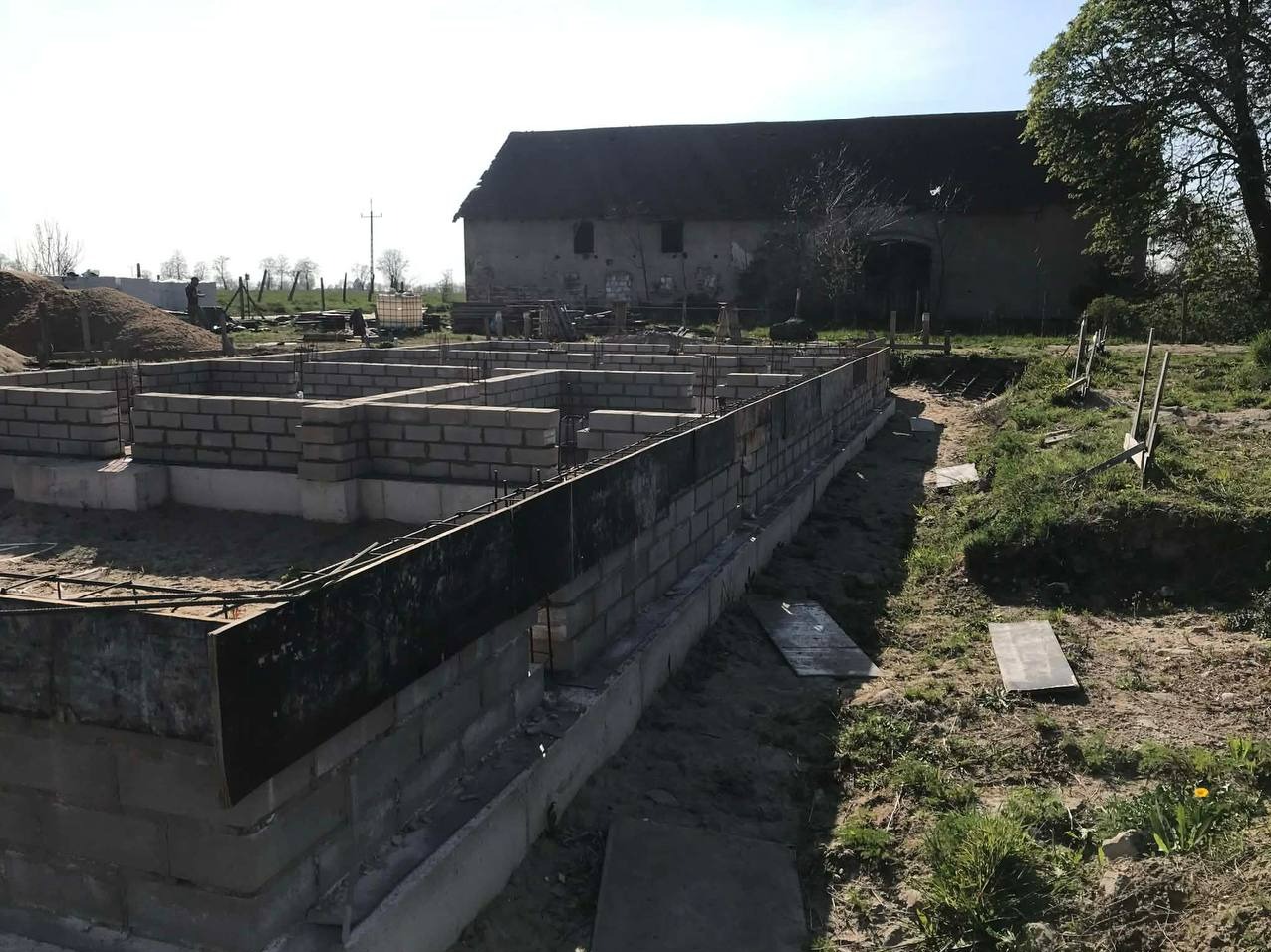 Wieniec fundamentowy z szalunkiem i zbrojeniem na budowie domu jednorodzinnego. Widoczne bloczki betonowe, stalowe pręty i deski szalunkowe. W tle budynek gospodarczy.