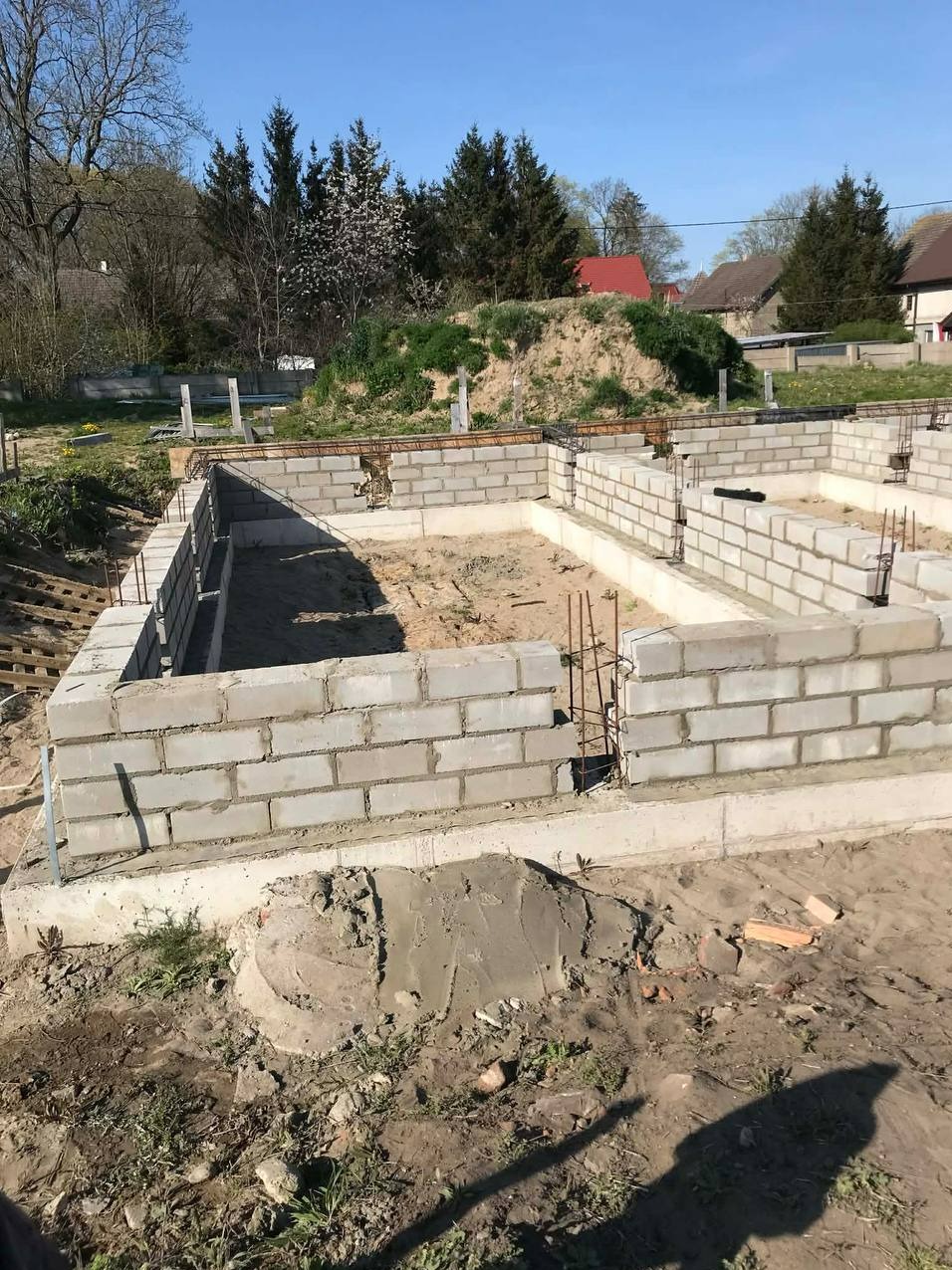 Fundament domu w trakcie budowy: ściany z bloczków betonowych, zbrojenie, piasek wewnątrz. Widok z góry, słoneczny dzień, domy w tle.