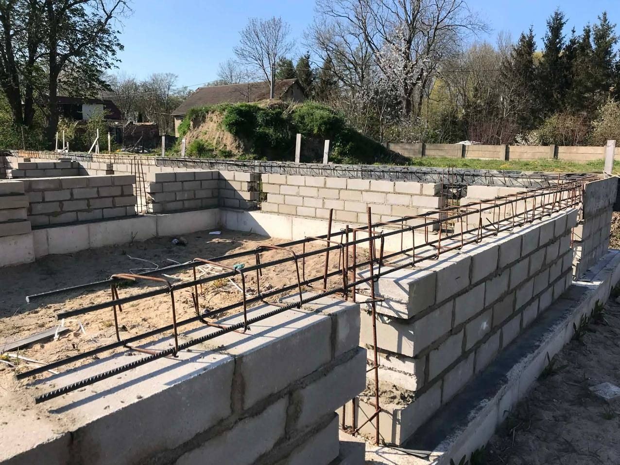 Zbrojenie wieńca fundamentowego z szalunkiem z bloczków betonowych na tle zieleni i zabudowań. Widok na konstrukcję fundamentów domu w trakcie budowy.
