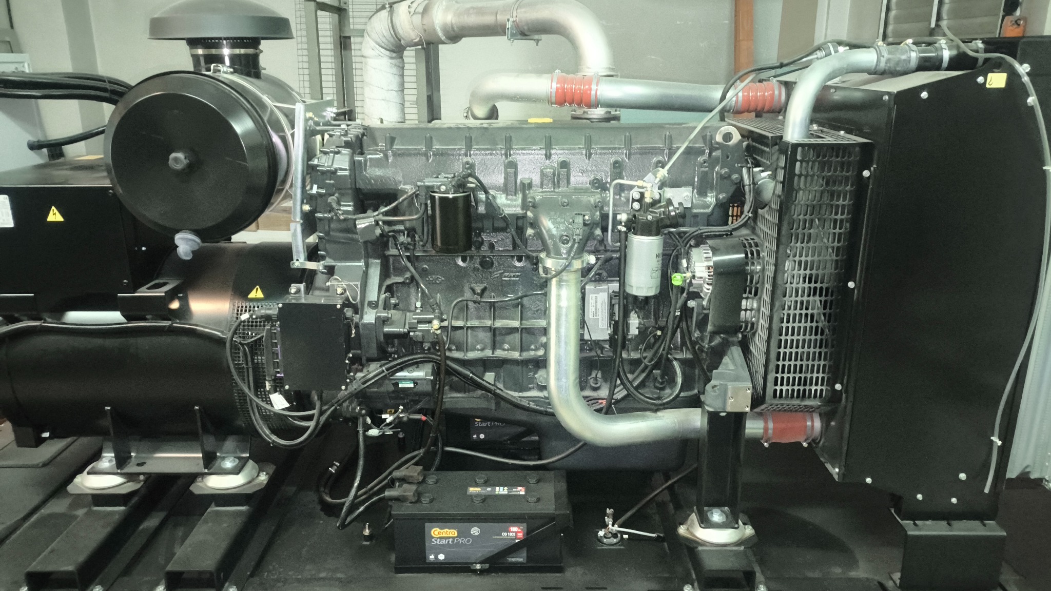 Czarny agregat prądotwórczy z widocznymi przewodami, filtrami i akumulatorem Centra Start PRO, umieszczony na metalowej konstrukcji w hali.