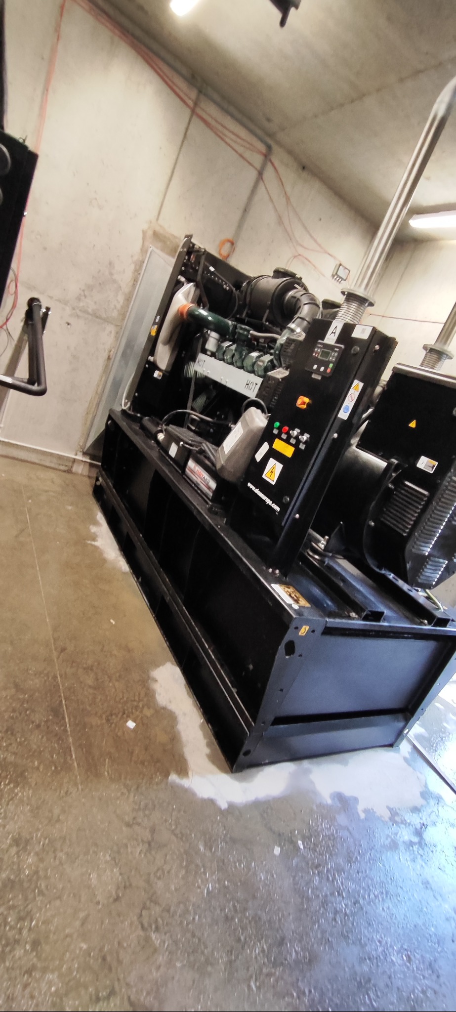 Czarny agregat prądotwórczy w pomieszczeniu z betonowymi ścianami i podłogą. Widoczne elementy silnika, panel sterowania i tabliczki ostrzegawcze.