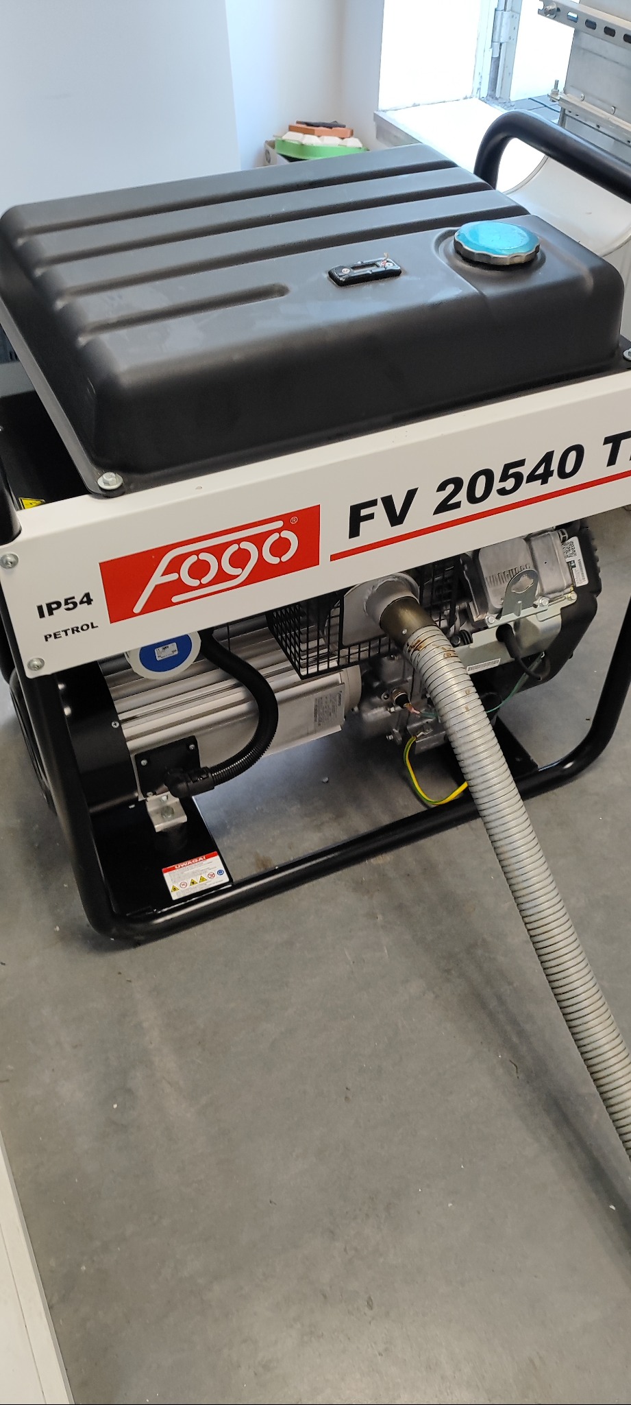 Agregat prądotwórczy Fogo FV 20540 T z widoczną tabliczką znamionową, umieszczony na szarej posadzce. Widoczny przewód odprowadzający spaliny.