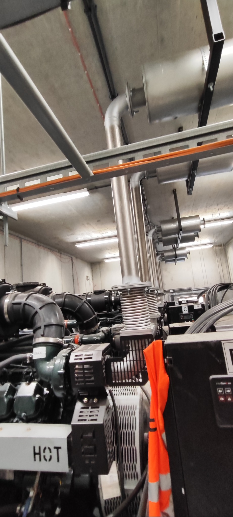 Agregat prądotwórczy w pomieszczeniu technicznym z widocznymi rurami wydechowymi i elementami instalacji. Na pierwszym planie fragment pomarańczowej kamizelki.