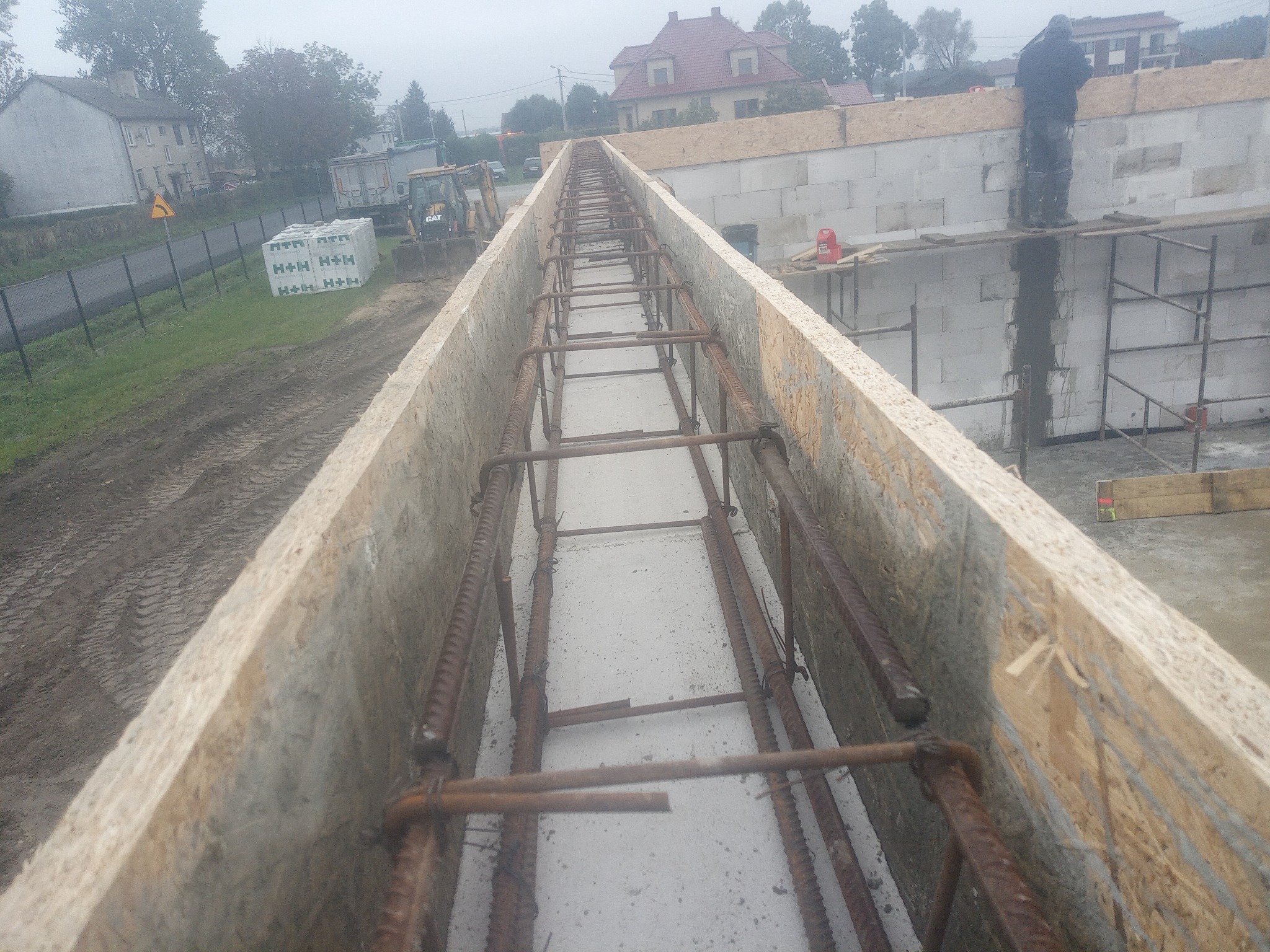 Zbrojenie fundamentów w szalunku z desek, widok z góry. W tle budynek mieszkalny i maszyny budowlane. Prace fundamentowe w toku, widoczny pracownik na rusztowaniu.