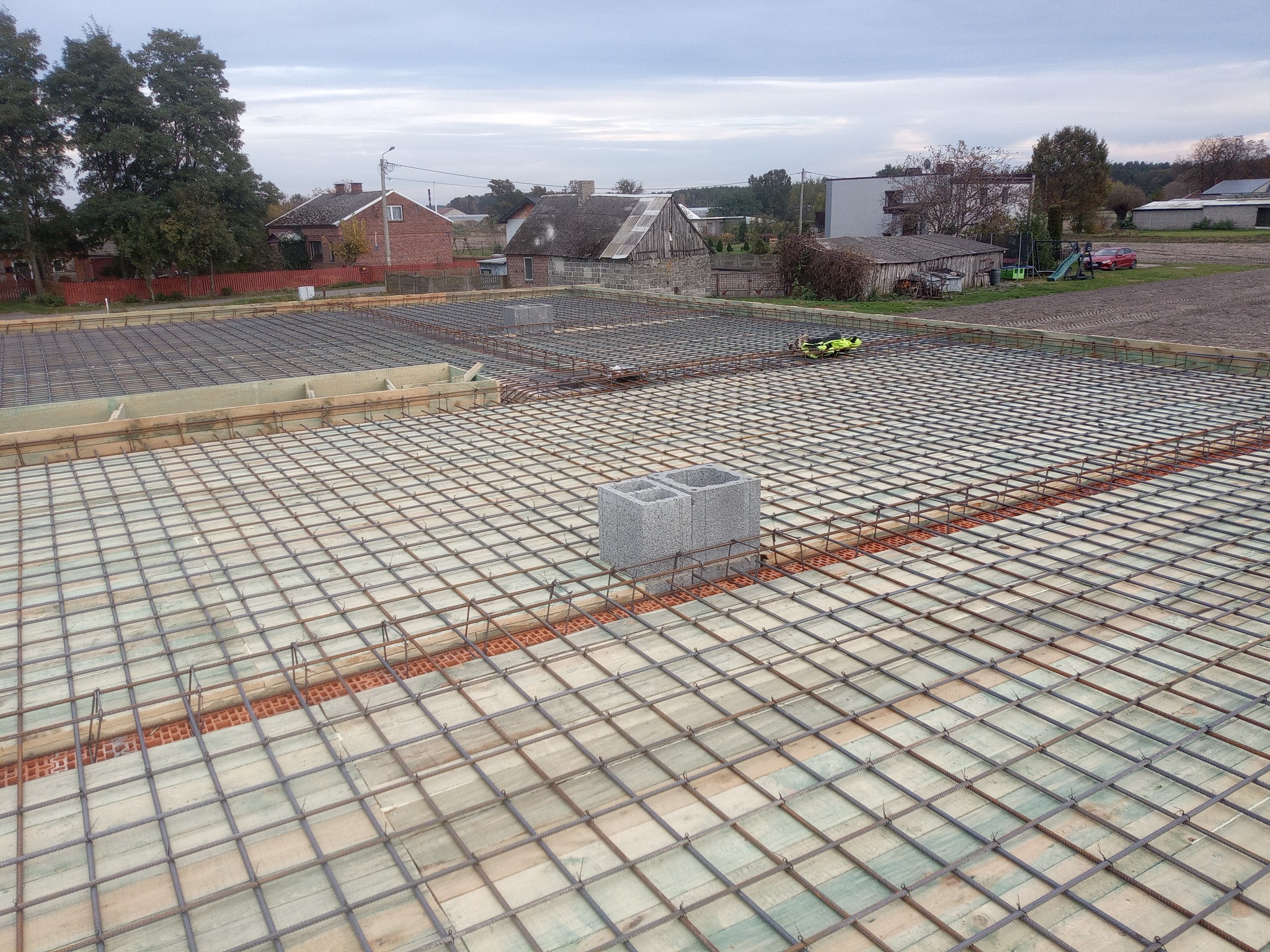 Zbrojenie fundamentów domu jednorodzinnego z widocznymi bloczkami betonowymi i deskowaniem. W tle zabudowa wiejska pod pochmurnym niebem.