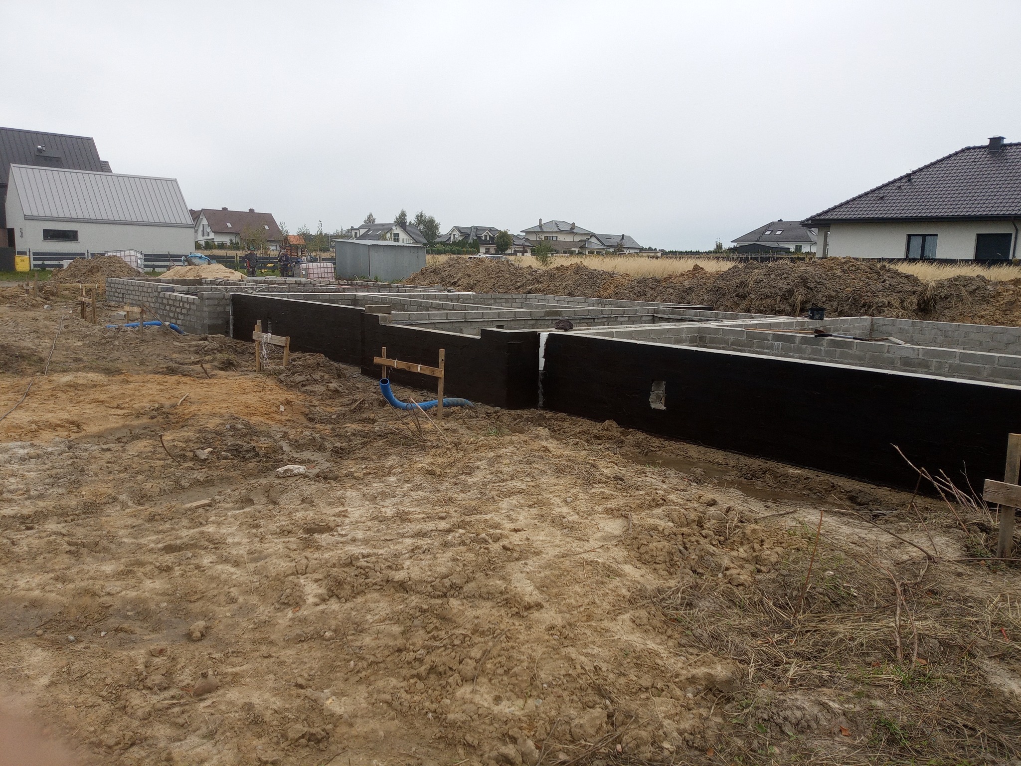 Fundamenty domu jednorodzinnego w trakcie budowy, z widoczną izolacją przeciwwilgociową i częściowo wymurowanymi ścianami z bloczków betonowych. Teren budowy z ziemią.