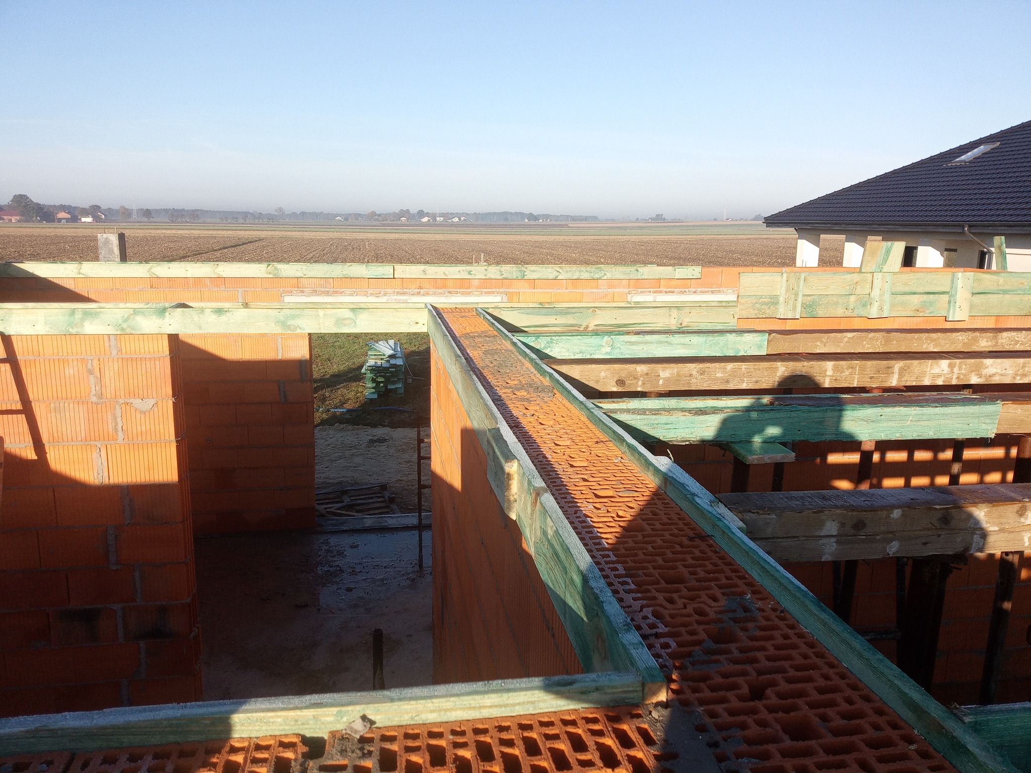 Wznoszenie ścian domu z pustaków ceramicznych, widok z góry na budowę w trakcie realizacji, z zielonymi elementami szalunku i widokiem na pola w tle.