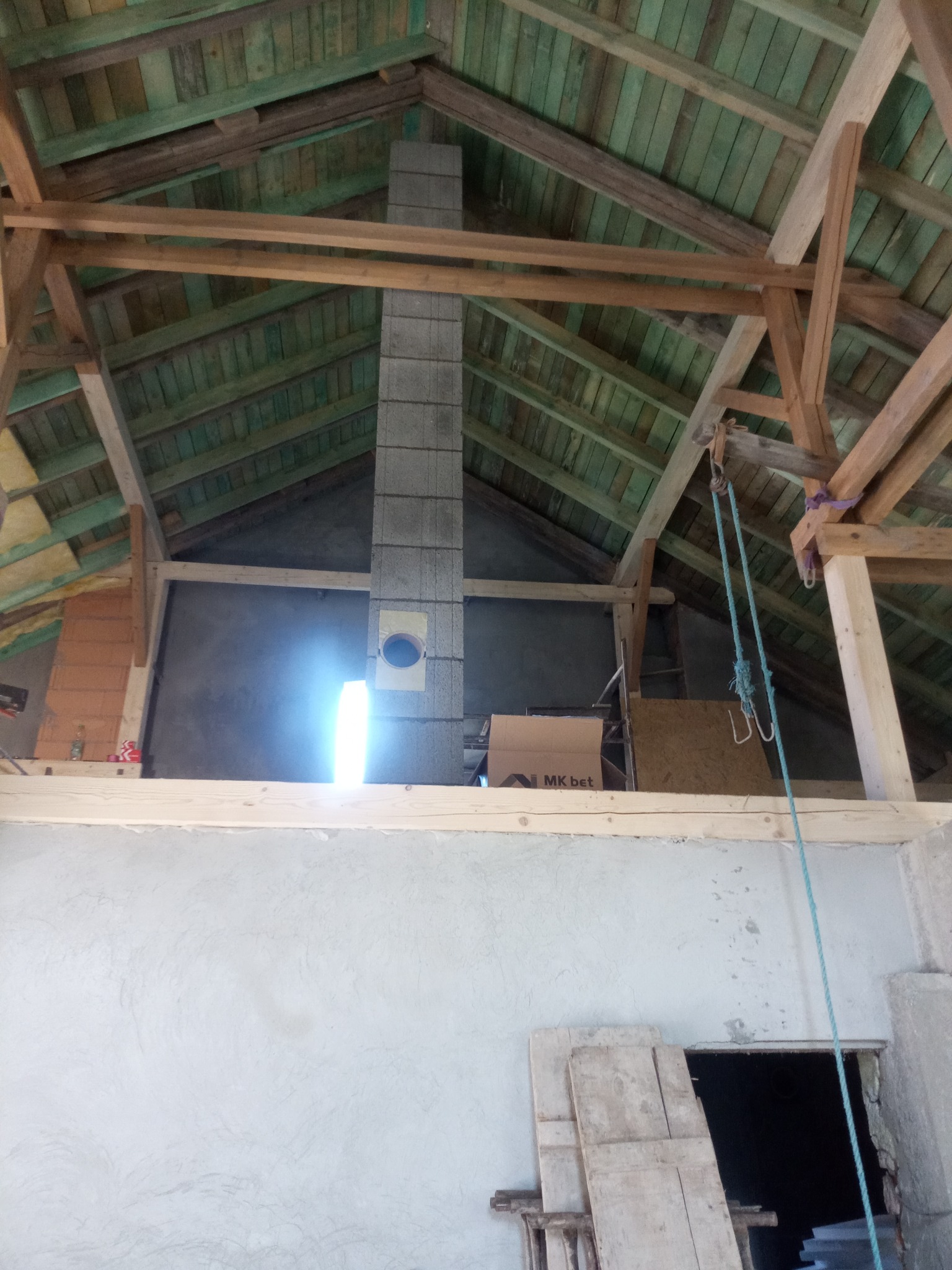 Wnętrze w trakcie budowy: widoczny komin z szarej cegły, drewniana konstrukcja dachu z zielonymi deskami, szara ściana i lina. Ujęcie z dołu, ukazujące postęp prac.