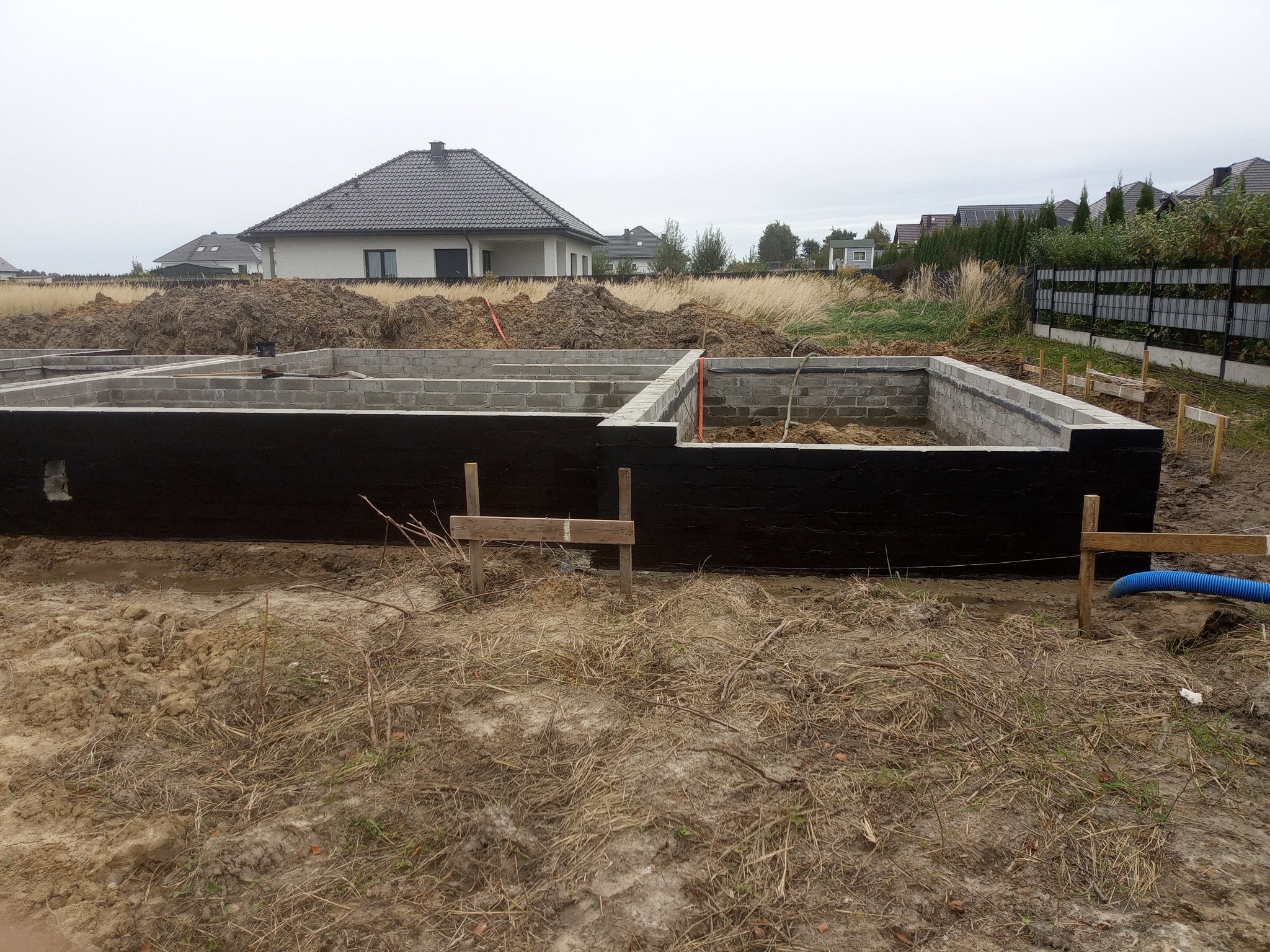 Zabezpieczone fundamenty z bloczków betonowych, izolowane czarną masą bitumiczną. W tle dom w zabudowie jednorodzinnej i przygotowany teren pod budowę.