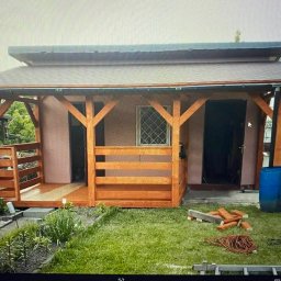 M&W Transport - Drewniana altana z zadaszeniem, częściowo osłonięta balustradą, z widocznymi elementami konstrukcyjnymi i oknem z kratką, otoczona zielenią i elementami budowlanymi.