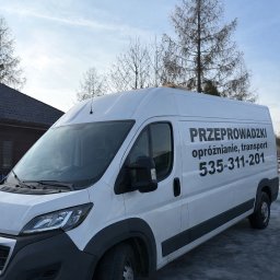 M&W Transport - Biały bus z napisem 'Przeprowadzki, opróżnianie, transport' i numerem telefonu na boku, zaparkowany na zewnątrz, w tle budynek i drzewa bez liści.