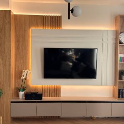 ProInstal Petro Hantsiuk - Nowoczesna zabudowa TV z drewnianymi panelami i podświetleniem LED, uzupełniona o półki i szafki w odcieniach beżu. Minimalistyczny design z akcentami roślinnymi.