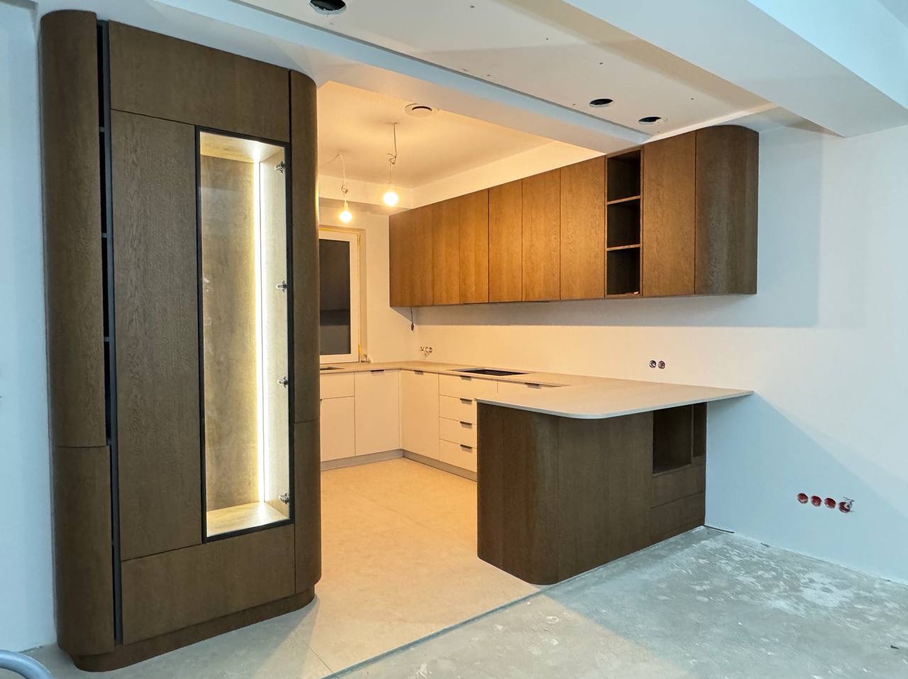 Nowoczesna kuchnia z zaokrąglonymi szafkami z drewna w odcieniach brązu i bieli, oświetlona listwą LED w witrynie. Widoczne gniazdka elektryczne na ścianie.