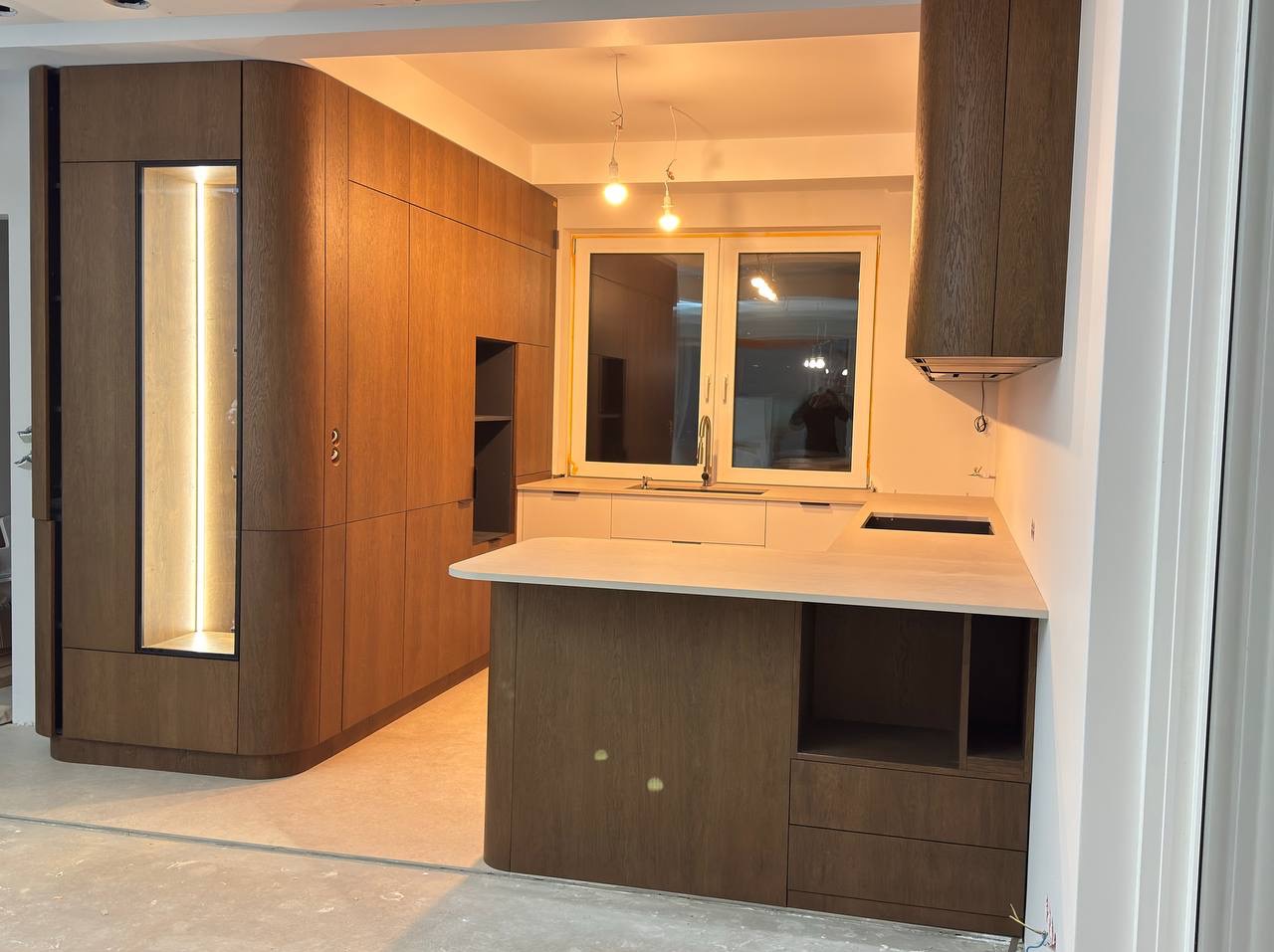 Nowoczesna kuchnia z drewnianymi, zaokrąglonymi szafkami, wyspą i oświetleniem LED. Minimalistyczny design z jasnymi blatami i dużym oknem.