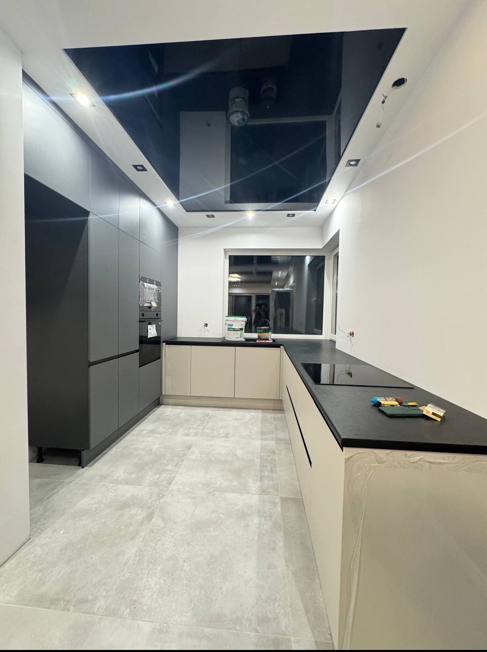 Nowoczesna kuchnia z czarnymi i beżowymi szafkami, czarnym sufitem podwieszanym i oknem w tle. Na blacie widoczne akcesoria malarskie.