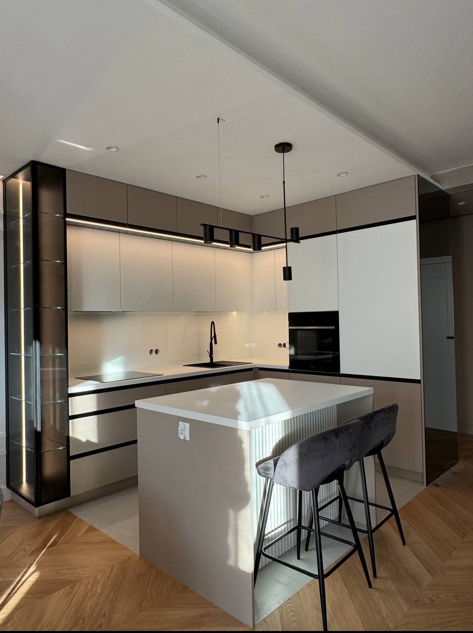 Nowoczesna kuchnia w beżach i bieli z wyspą, czarnymi akcentami oświetlenia i hokerami. Parkiet w jodełkę. Szklana witryna z podświetleniem.