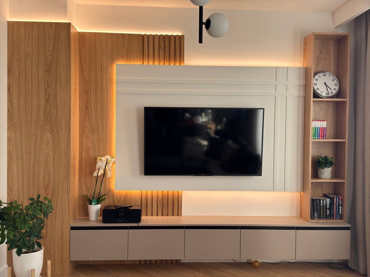 Nowoczesna zabudowa TV z drewnianymi panelami i podświetleniem LED, uzupełniona o półki i szafki w odcieniach beżu. Minimalistyczny design z akcentami roślinnymi.