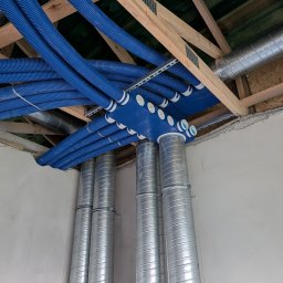 Jakub Siejda - Niebieskie, elastyczne przewody wentylacyjne pod stropem, połączone z metalowymi rurami. Widoczne drewniane elementy konstrukcyjne i fragment ściany. Instalacja w trakcie montażu.