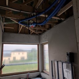 Jakub Siejda - Niebieskie rury wentylacyjne pod drewnianym stropem w surowym wnętrzu. Widoczne okna z folią ochronną i stelaż podtynkowy WC Grohe. Etap montażu instalacji.