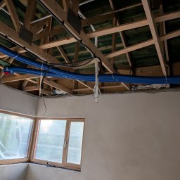 Jakub Siejda - Montaż instalacji wentylacyjnej z niebieskimi rurami w surowym wnętrzu. Widoczne drewniane elementy konstrukcyjne dachu i okno z folią ochronną.