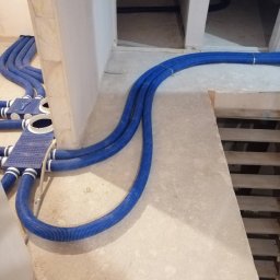 Jakub Siejda - Niebieskie, karbowane rury wentylacyjne ułożone w surowym wnętrzu, z widocznymi elementami montażowymi i fragmentem schodów. Instalacja rekuperacji w trakcie prac.