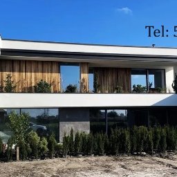 TERMI Elewacje - Nowoczesny dom z białą elewacją i drewnianymi panelami na balkonie. Widoczne duże okna i starannie zagospodarowany teren wokół budynku. Na górze zdjęcia numer telefonu.