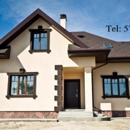 TERMI Elewacje - Elewacja domu jednorodzinnego w jasnym odcieniu z brązowymi detalami: okna, dach, obramowania. Widoczny numer telefonu. Ujęcie z poziomu gruntu, błękitne niebo w tle.