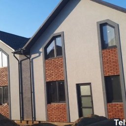 TERMI Elewacje - Dwukondygnacyjny dom z szarą elewacją i ceglanymi wstawkami wokół okien. Widoczne rynny i fragment dachu. Na pierwszym planie hałda piasku.