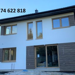 TERMI Elewacje - Dwukondygnacyjny dom z białą elewacją i drewnianymi panelami. Widoczne okna z drewnianymi ramami i duże przesuwne drzwi tarasowe. Na górze numer telefonu.