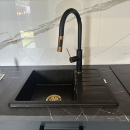 Ivan Lukashuk - Czarny, granitowy zlew z ociekaczem i złotym odpływem, bateria w kolorze czarnym z elementami złota, marmurowa ściana w tle. Nowoczesna kuchnia.