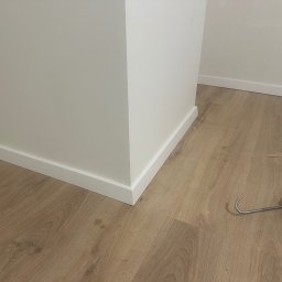 Ivan Lukashuk - Wykończona podłoga z panelami i listwami przypodłogowymi przy ścianie. Widoczny aplikator do silikonu i resztki materiałów.