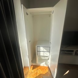 Ivan Lukashuk - Otwarta, biała szafa z półkami i drążkiem na ubrania, widoczna na tle parkietu i fragmentu zasłony. Szafa oświetlona naturalnym światłem.