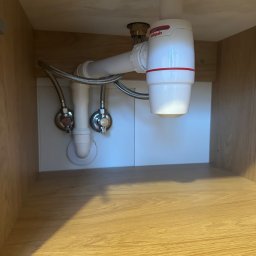 Ivan Lukashuk - Syfon umywalkowy z białego tworzywa sztucznego, zamontowany w szafce z drewnianymi ściankami. Widoczne wężyki doprowadzające wodę i zawory.