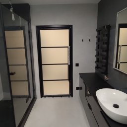 A.R.M. - Minimalistyczna łazienka w szarościach z czarną kabiną prysznicową i drzwiami z matowymi panelami. Biała umywalka nablatowa na ciemnej szafce, obok czarny grzejnik. Widoczna wisząca żarówka.