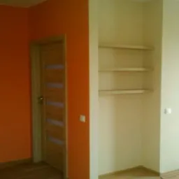 Wnętrze pokoju po remoncie: pomarańczowa ściana, drewniane drzwi z przeszkleniami, wnęka z półkami i podłoga z paneli.