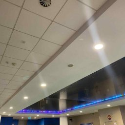 Bartosz Turek - Sufit podwieszany z oświetleniem LED w nowoczesnym biurze. Widoczne panele sufitowe, okrągłe lampy i czarny element dekoracyjny z niebieskim podświetleniem.