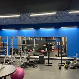 Bartosz Turek - Nowoczesna siłownia z niebieskimi ścianami, lustrem, sprzętem do ćwiczeń i piłką fitness. Widoczne oświetlenie sufitowe i gumowa podłoga.