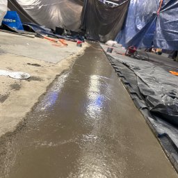 Bartosz Turek - Wyrównywanie podłoża wylewką samopoziomującą, widoczne narzędzia i zabezpieczenia. Prace wykończeniowe w pomieszczeniu z szarym podłożem i folią ochronną.