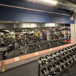 Bartosz Turek - Wnętrze siłowni z rzędami hantli, maszynami do ćwiczeń i dużym lustrem. Widoczny mężczyzna z telefonem i odkurzacz, co sugeruje sprzątanie lub prace porządkowe.