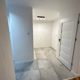 Bartosz Turek - Jasne wnętrze po remoncie: szare płytki podłogowe, białe ściany i drzwi. Widoczne detale wykończenia, minimalistyczne oświetlenie sufitowe. Nowoczesny design.