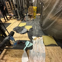 Bartosz Turek - Remont siłowni w toku: podłoga z płyt OSB, zakryte folią sprzęty, narzędzia i butelka wody. Widoczne prace przygotowawcze, prawdopodobnie przed malowaniem.