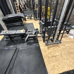 Bartosz Turek - Sprzęt do ćwiczeń siłowych w siłowni, z czarnymi matami podłogowymi i jasną podłogą z płyty OSB. Widoczny fragment lustra i osoba w tle.