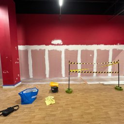 Bartosz Turek - Wnętrze w trakcie prac: czerwona ściana z pasami szpachli, narzędzia malarskie, taśma ostrzegawcza. Podłoga wyłożona panelami imitującymi drewno.