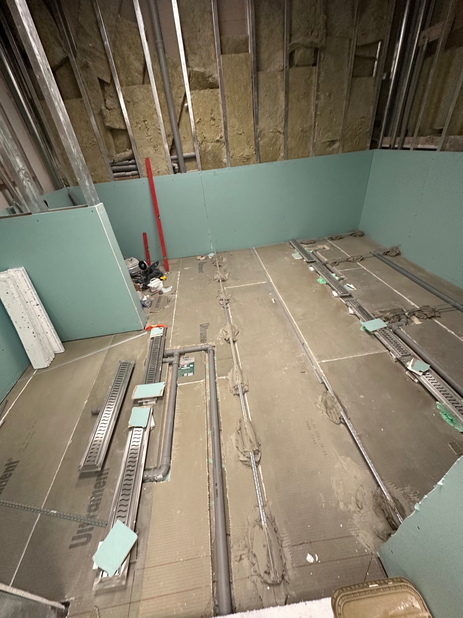 Montaż odpływów liniowych w łazience na poddaszu. Widoczne płyty podłogowe, rury odpływowe i profile. Ściany wykończone płytami gipsowo-kartonowymi, ocieplenie wełną mineralną.